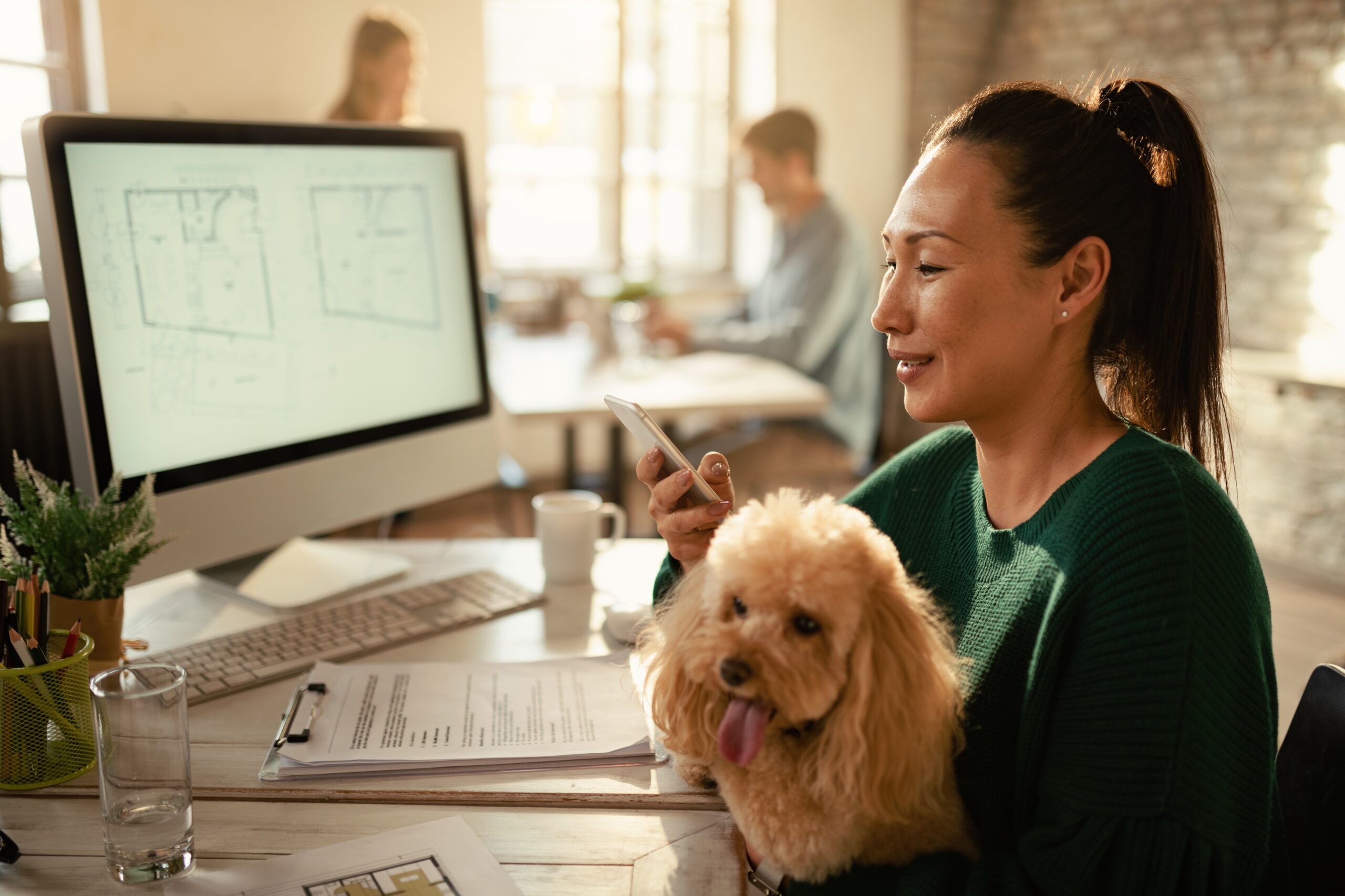Smiling Asian entrepreneur using smart phone while holding her poodle in the office.